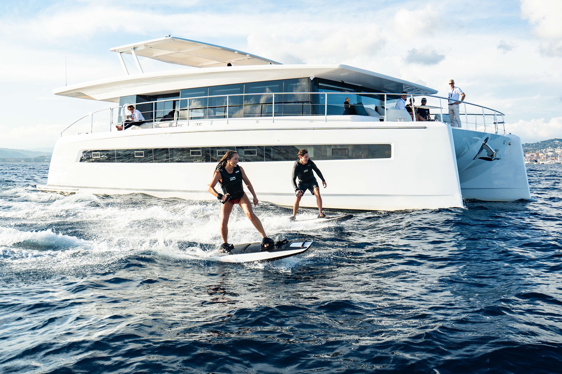 A man and a woman ride Awake RÄVIK jetboards, effortlessly speeding across the water with an electric yacht in the background, capturing a moment of excitement and luxury.